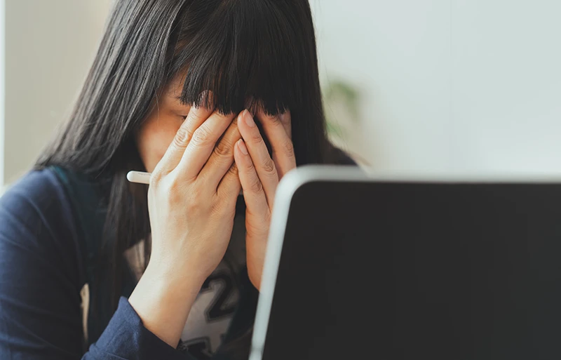 a young woman covering her eyes with her hands while using a laptop